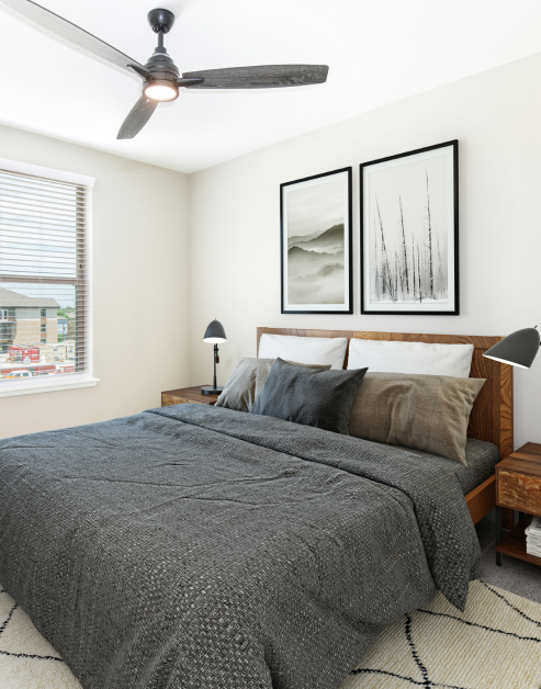 Model bedroom with wall pictures above grey bed with pillows, ceiling fan and wood nightstands with lamps next to a window with blinds at MAA Park Point luxury apartments in Houston, TX