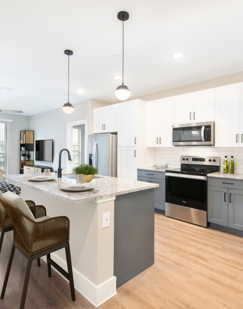 Open-style modern kitchen with white cabinets with tiled backsplash and stainless steel appliances, hanging light fixtures over island countertop with sink and bar chairs on light wood floors at MAA Park Point luxury apartments in Houston, TX