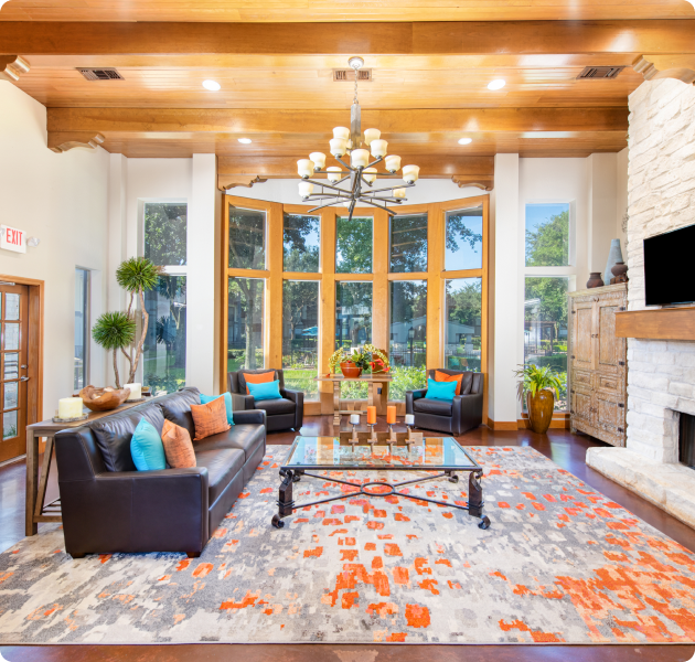 Community clubhouse with wood beam ceiling and bay windows with a chandelier, lounge sitting area on colorful area rug next to brick fireplace and wall-mounted TV at MAA Ranchstone luxury apartments in Houston, TX
