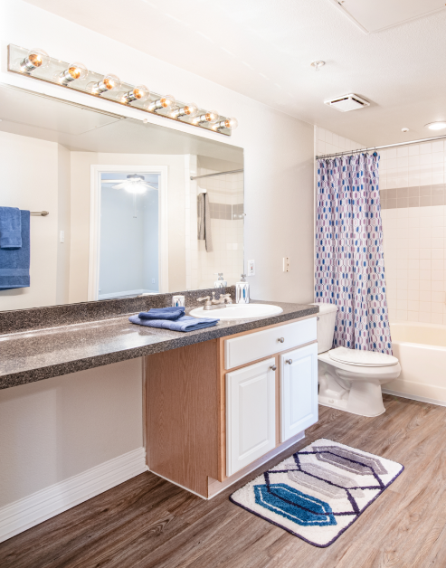 Bathroom with light fixtures above a large vanity mirror, countertop with sink and wood cabinets next to shower curtains on wood floors at MAA Ranchstone luxury apartments in Houston, TX