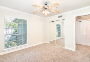 Empty carpeted bedroom with ceiling fan, mirrored sliding doors next to the large picture window with blinds at MAA Ranchstone luxury apartments in Houston, TX