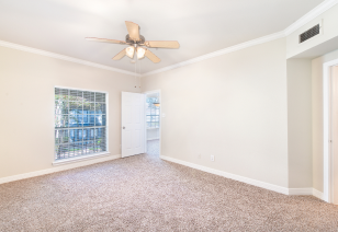 Empty carpeted bedroom with ceiling fan and large picture widow with blinds next to an opened white door at MAA Ranchstone luxury apartments in Houston, TX