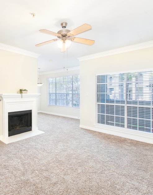 Empty carpeted living room with a ceiling fan, fireplace and large picture windows with blinds at MAA Ranchstone luxury apartments in Houston, TX