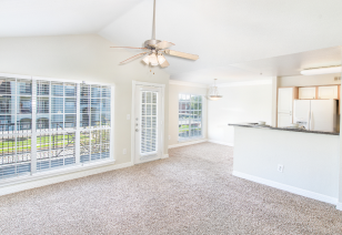 Empty carpeted living room with vaulted ceiling and hanging fan, a French door and large picture windows with blinds next to the kitchen bar top counter at MAA Ranchstone luxury apartments in Houston, TX