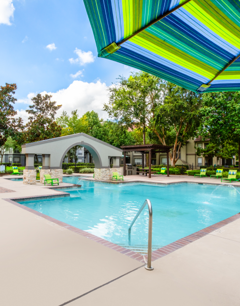 Long view of resort-style pool with entrance steps and a metal handrail surrounded by green lounge chairs, a shaded wooden pergola and lush trees at MAA Ranchstone luxury apartments in Houston, TX