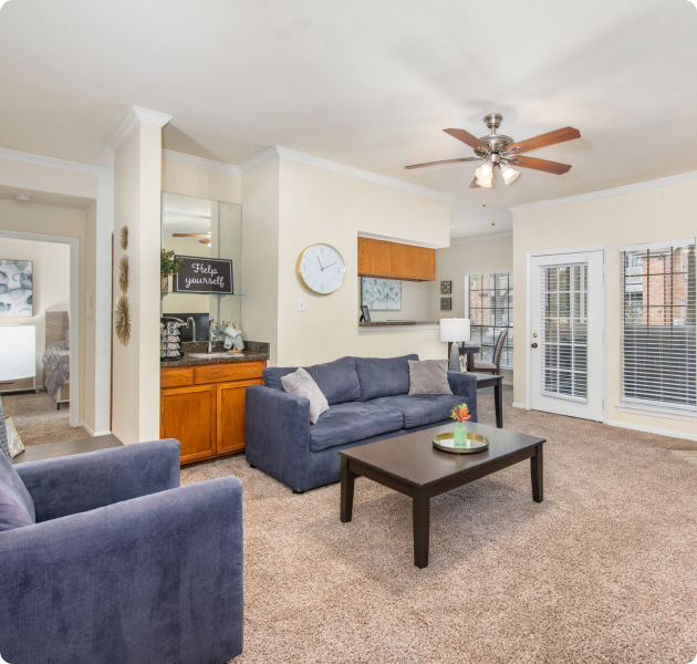 Apartment living area with ceiling fan, wall mounted clock next to a coffee bar and sitting area with wooden coffee table next to a French door and window with blinds at MAA Valleywood luxury apartments in the Woodlands, TX