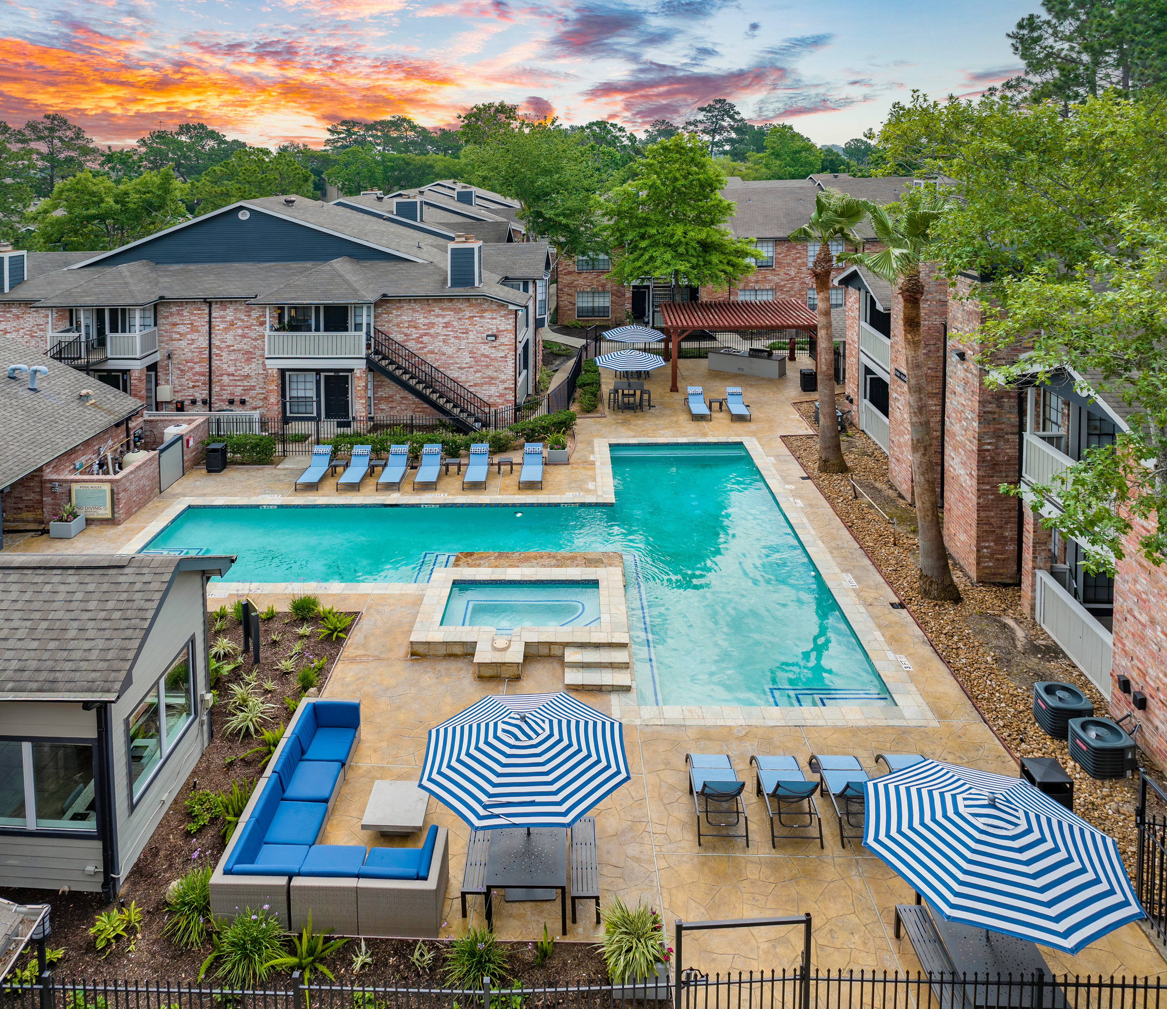 Aerial view of resort-style pool with lounge chairs, sun umbrellas with patio tables and pergola surrounded by lush trees at MAA Valleywood luxury apartments in the Woodlands, TX
