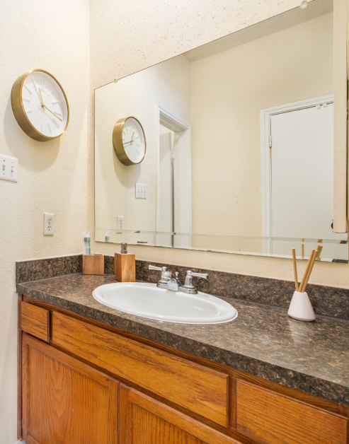 Modern bathroom with wall mounted clock, vanity mirror and sink on dark countertop with wood cabinets at MAA Valleywood luxury apartments in the Woodlands, TX