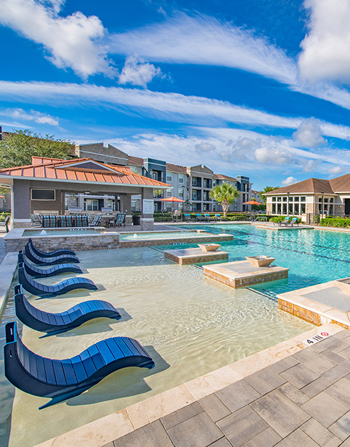 Side view of resort-style pool with modern sun deck, a covered patio area and lounge chairs surrounded by lush greenery next to the apartment complex at MAA Vintage Park luxury apartments in Houston, TX