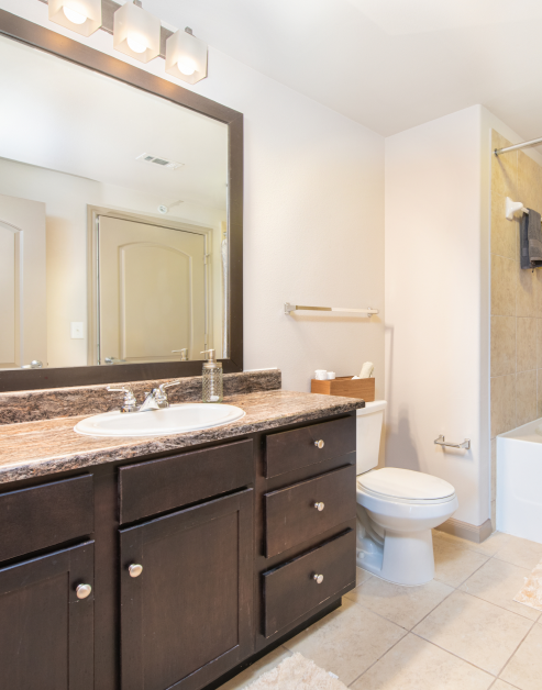 Modern-style bathroom with light fixtures above a dark wood framed vanity mirror, granite countertop and dark brown cabinets on tile floors at MAA Vintage Park luxury apartments in Houston, TX