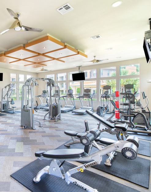 A spacious fitness center with ceiling fans, wall mounted TVs, treadmills and a variety of workout equipment next to large windows on tile flooring at MAA Vintage Park luxury apartments in Houston, TX