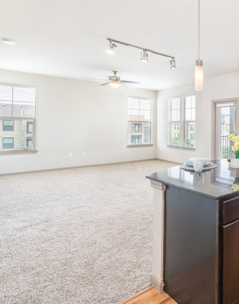 Spacious empty carpeted living room with a ceiling fan and windows with blinds next to kitchen area at MAA Vintage Park luxury apartments in Houston, TX