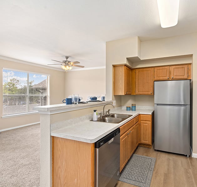 Open-style apartment kitchen with wood cabinets, white counter with stainless steel appliances on wood floors next to living area at MAA Woodwind luxury apartments in Houston, TX