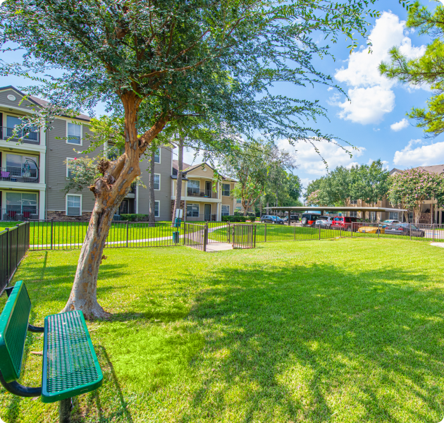 Community gated dog park with lush grass and trees and a green bench, enclosed with iron fence next to apartment complex at MAA Woodwind luxury apartments in Houston, TX