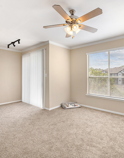 Empty carpeted living room with a ceiling fan and track light fixtures, large picture windows with blinds next to sliding glass doors at MAA Woodwind luxury apartments in Houston, TX