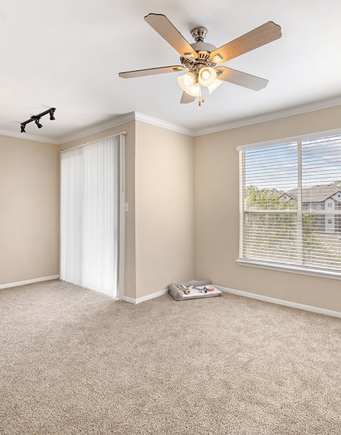 Empty carpeted living room with a ceiling fan and track light fixtures, large picture windows with blinds next to sliding glass doors at MAA Woodwind luxury apartments in Houston, TX
