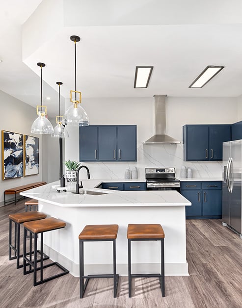 Clubhouse modern kitchen with hanging light fixtures over white bar top counter with a sink, dark blue cabinets, stainless steel appliances and bar stools on wood floors at MAA Woodwind luxury apartments in Houston, TX