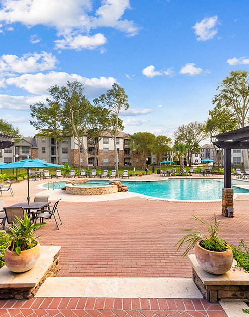 Long view of resort-style pool area with sundeck, sun umbrellas with patio tables, lounge chairs and a pergola surrounded by lush greenery at MAA Woodwind luxury apartments in Houston, TX
