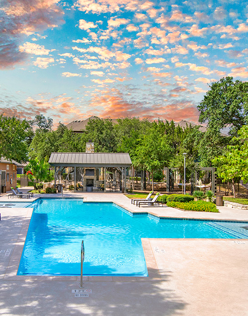 Resort-style pool with entrance steps and lounge chairs surrounded by lush trees situated next to large pergola at MAA Bulverde luxury apartments in San Antonio, TX