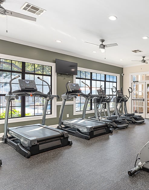 Fitness center with ceiling fans, wall mounted TV and row of treadmills next to large picture windows at MAA Haven at Blanco luxury apartments in the San Antonio, TX