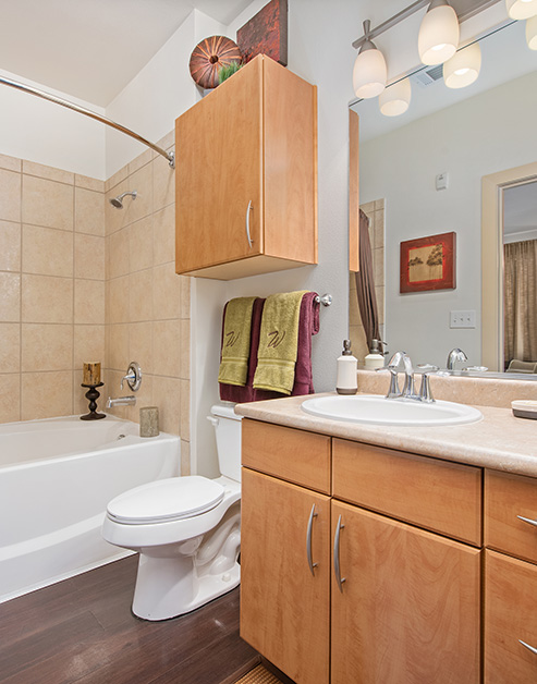 Bathroom with light fixtures above a large vanity mirror, light brown cabinets next to a bathtub with tiled walls on wood floor at MAA Westover Hills luxury apartments in San Antonio, TX