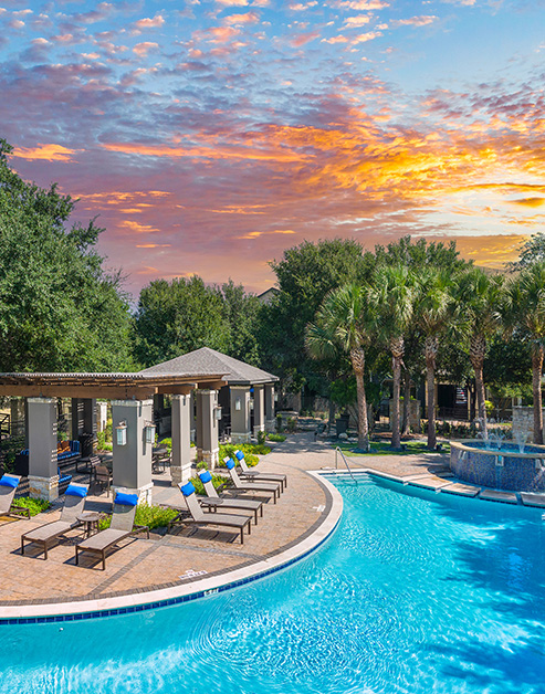 Pool with entrance steps, lounge chairs and pergola with multiple seating areas surrounded by lush trees with a sunset sky at MAA Westover Hills luxury apartments in San Antonio, TX
