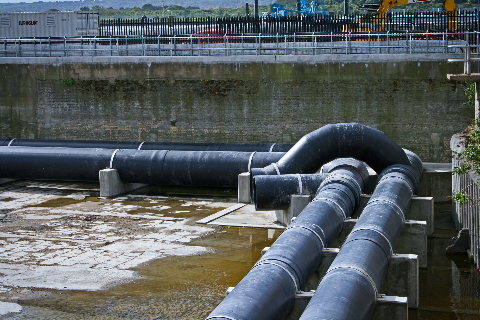 Image of thermoplastic piping in Beckton, with some lingering water on the ground and a concrete wall in the background