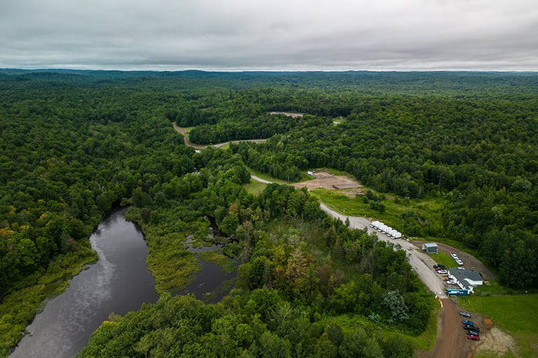 Aerial view of a forested landscape with a small water body, winding road, and a cluster of buildings