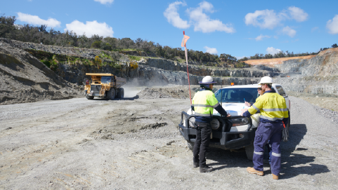 Three engineers standing around a car on bench in a quarry with a haul truck drving along a road in the background
