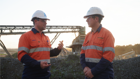 Two engineer's in white hard hats and orange high-visibility clothing conversing at a mine site.