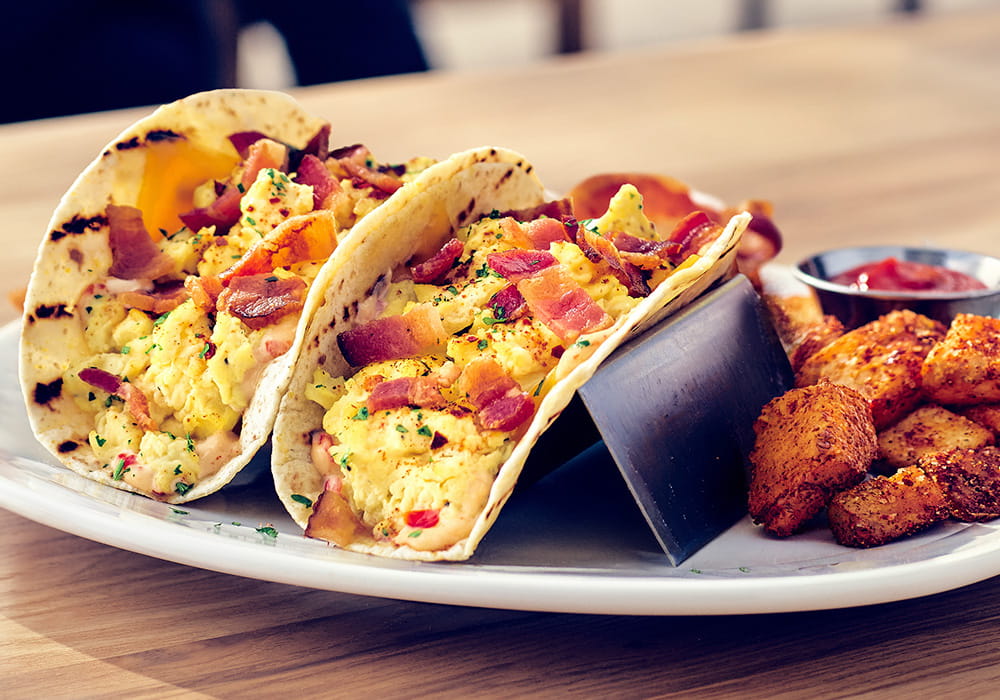 B.A.E. TACOS Crispy Bacon and cheesy scrambled Eggs, served in warm tortillas topped with cilantro, drizzled with our signature Bang Bang sauce, served with seasoned breakfast potatoes and applewood smoked bacon.