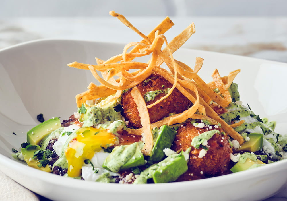 CRAB HUEVOS RANCHEROS Crispy crab bites, poached eggs, avocado and refried black beans, topped with enchilada sauce, cilantro-lime crema, feta cheese, and crispy tortilla strips.