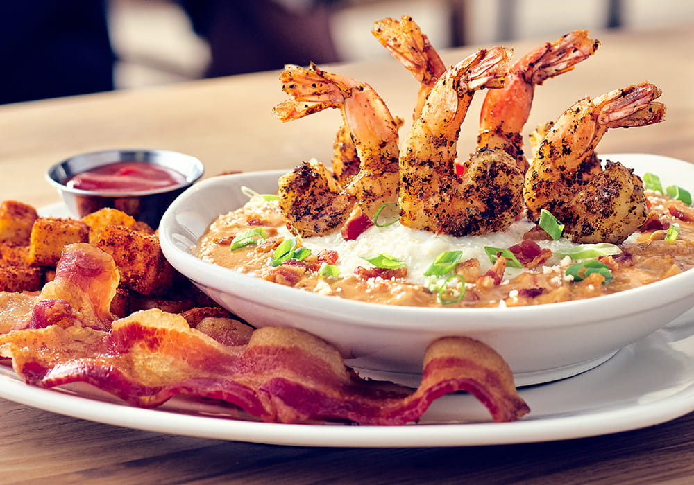 SHRIMP & GRITS Five wood-grilled shrimp topped with a creamy creole sauce, served over mozzarella cheese-infused grits, finished with green onions, parmesan cheese and bacon, served with seasoned potatoes and applewood smoked bacon.