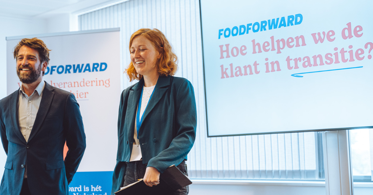 Twee personen bij een Food Forward presentatie, met banner en scherm waarop staat ‘Hoe helpen we de klant in transitie?’