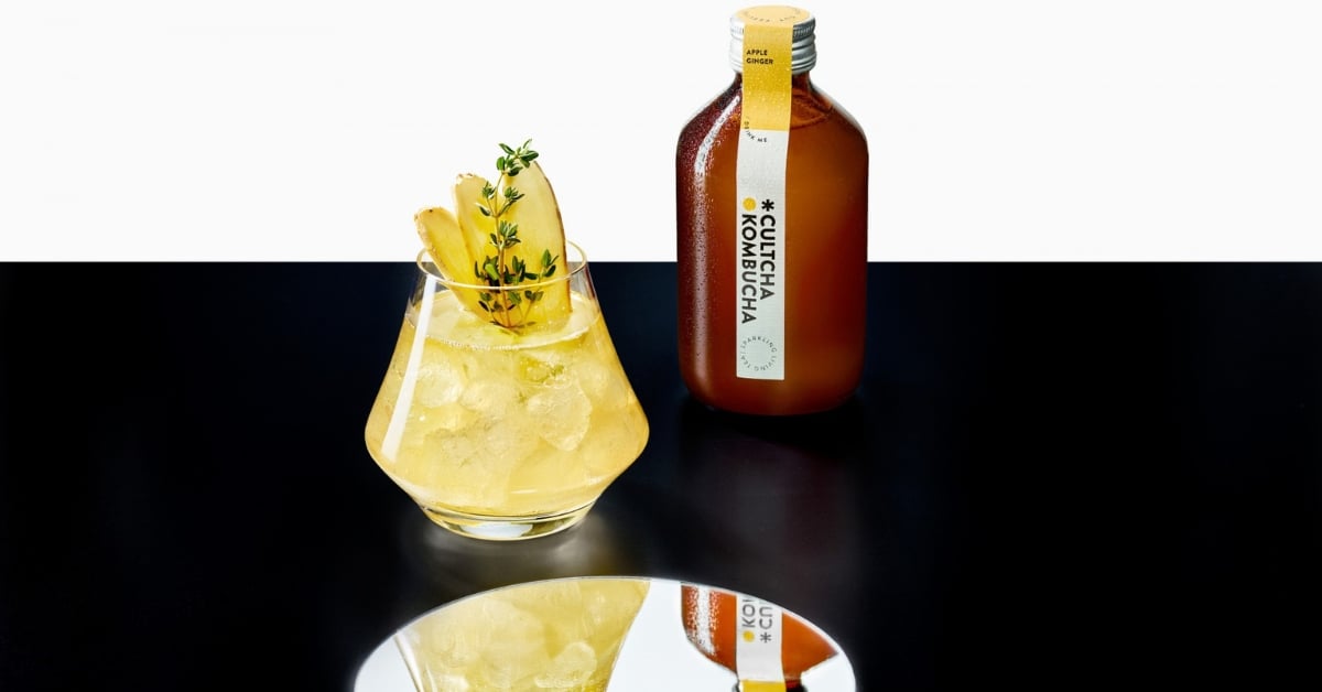 Glas met gele kombucha-cocktail met ijs, ananas en tijm, daarnaast staat een fles Cultcha Kombucha Apple Ginger