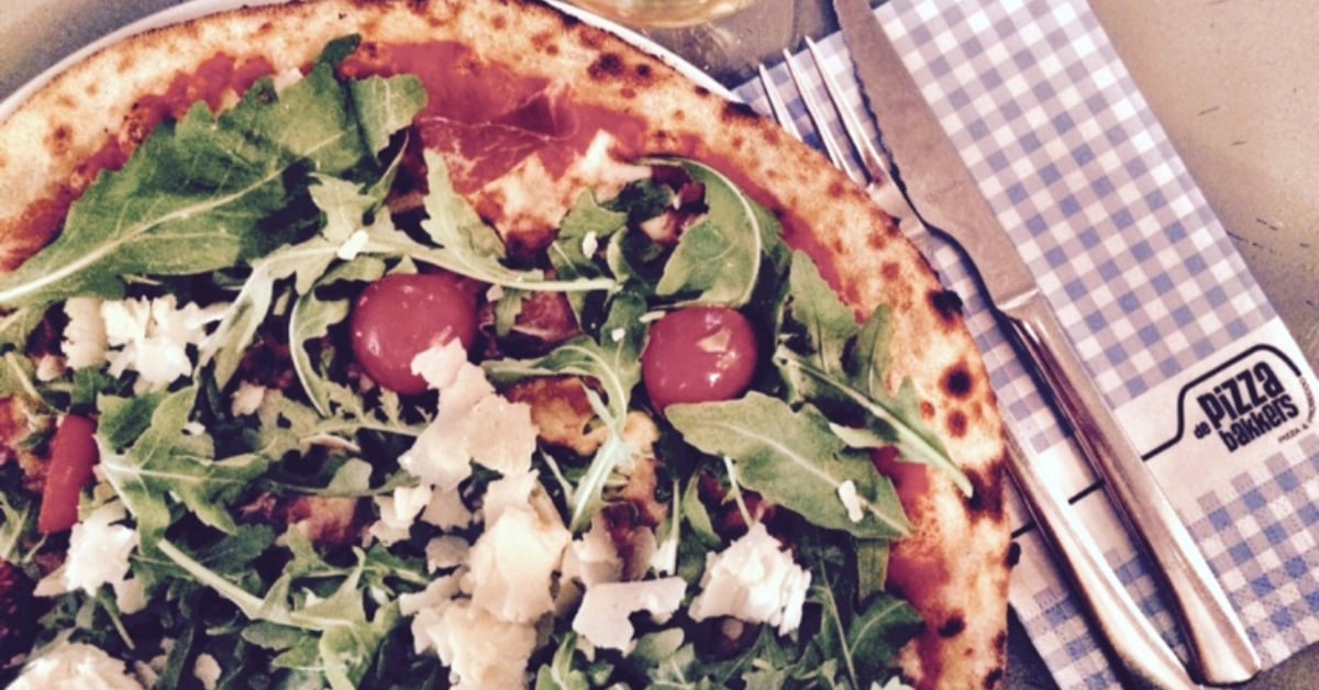 Close-up van een houtovenpizza van De Pizzabakkers met rucola, cherrytomaatjes en Parmezaanse kaas, geserveerd met bestek op geruit servet