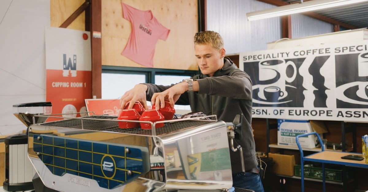Barista plaatst rode koffiekopjes op een espressomachine, met koffiebanners op de achtergrond
