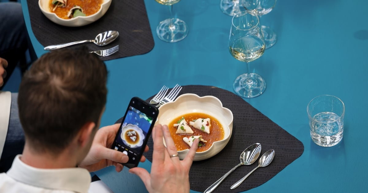 Man maakt foto van een opgemaakt gerecht in een kom met bestek en gazen witte wijn op tafel.