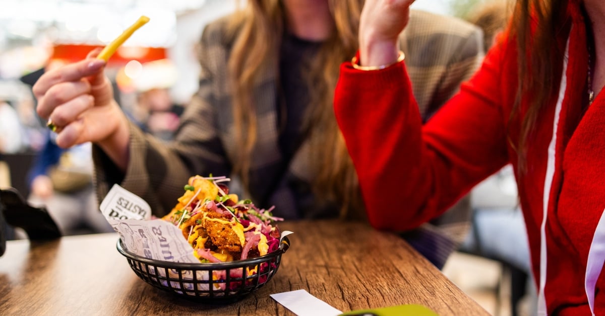 Twee mensen eten samen een bakje loaded friet aan een houten tafel.
