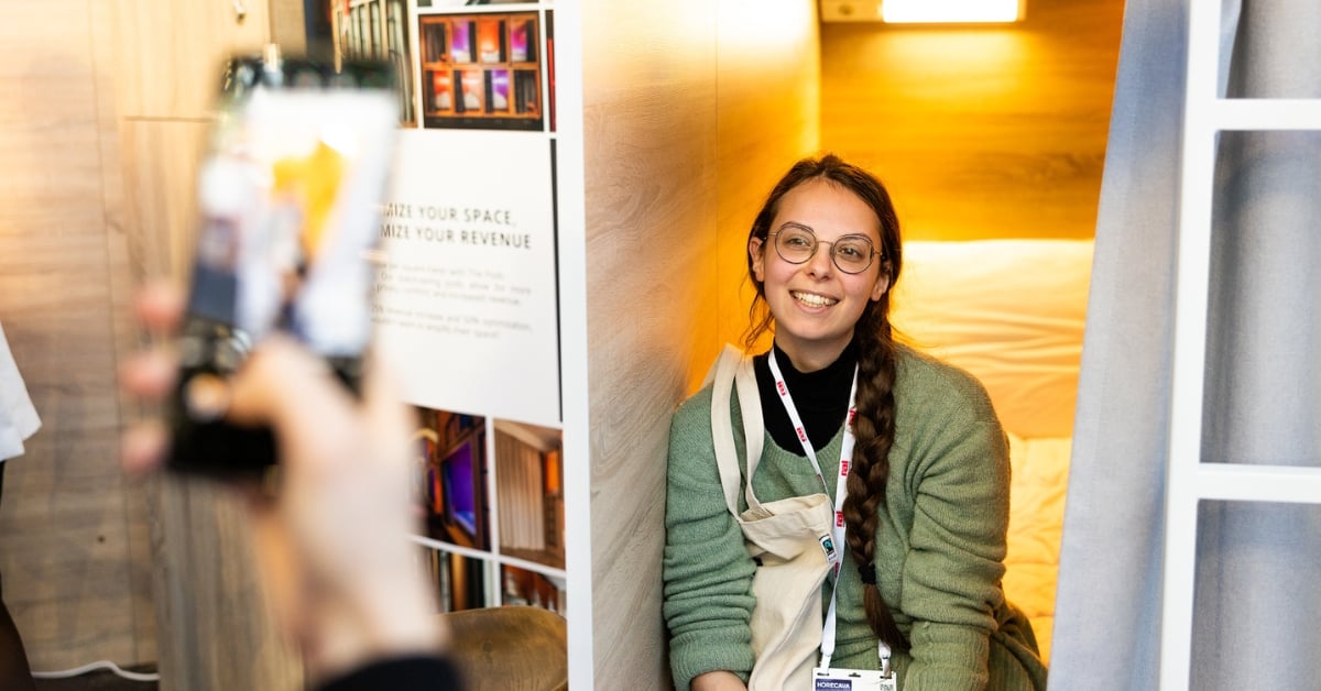 Vrouw met lange vlecht en bril lacht naar de camera, zittend in een compacte hotelruimte met warme verlichting.