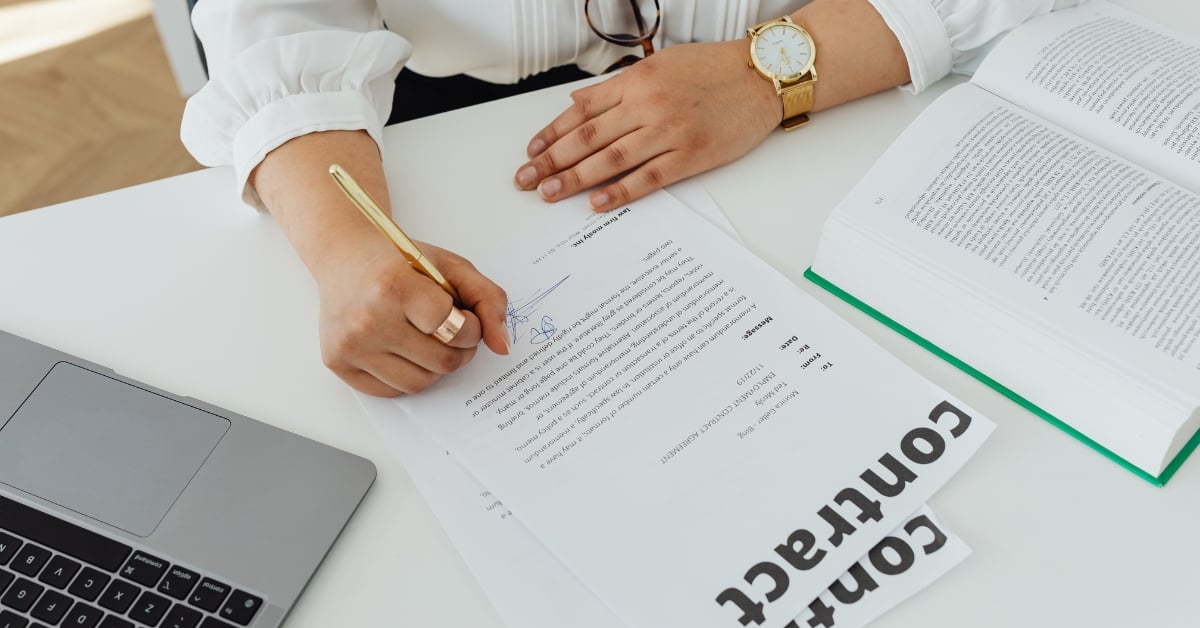 Close-up van handen die een contract ondertekenen naast een laptop en open boek.