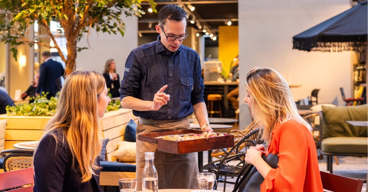 Gastheer in horecazaak toont assortiment aan twee vrouwelijke gasten op het terras