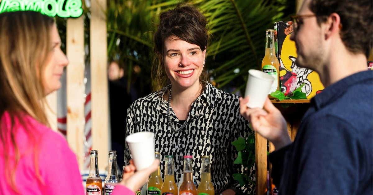 Vrouw achter een drankkraam praat met twee bezoekers die papieren bekers vasthouden
