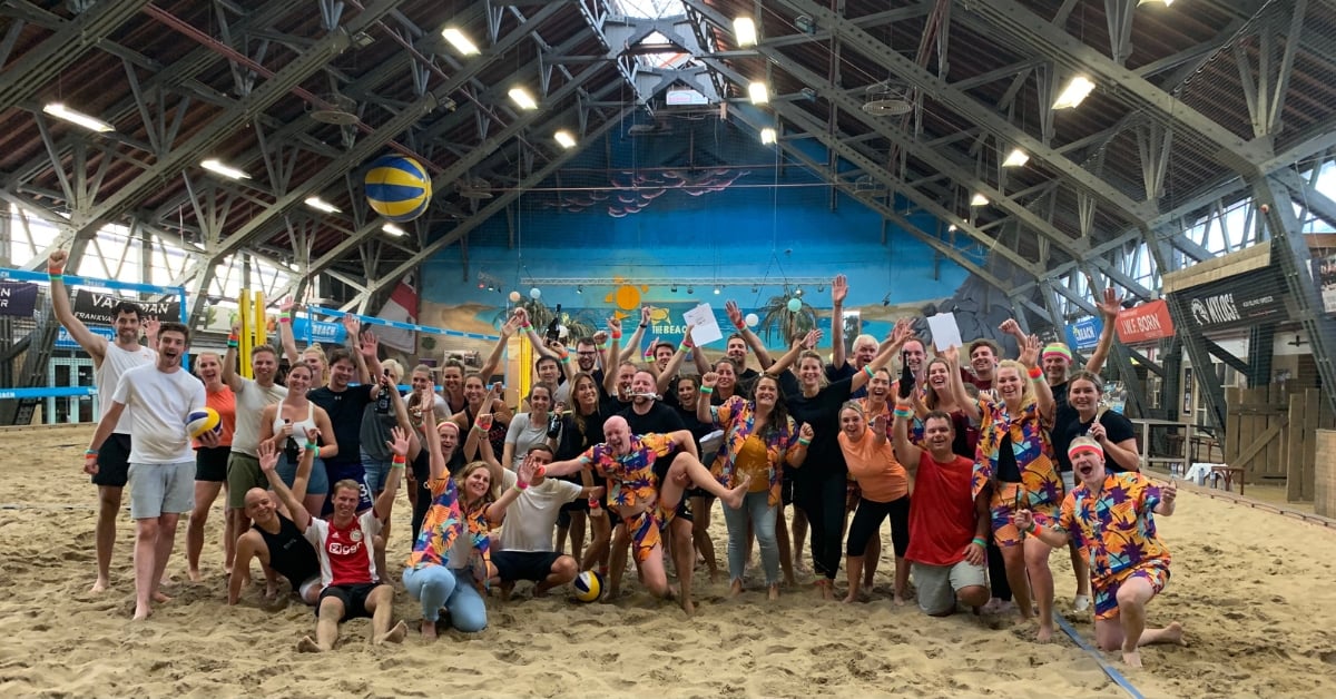 Grote groep collega's die vrolijk poseren op het indoorstrand van The Beach Aalsmeer tijdens een teamactiviteit, met volleybalnetten op de achtergrond.