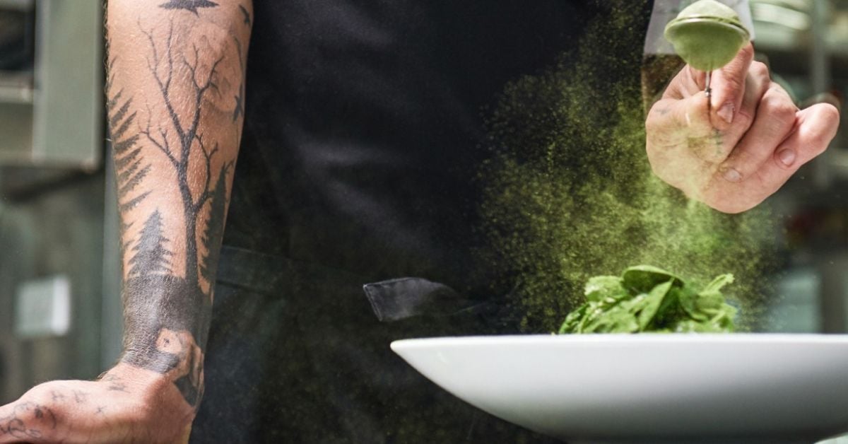 Chef strooit groen poeder over een gerecht met verse groene ingrediënten op een wit bord