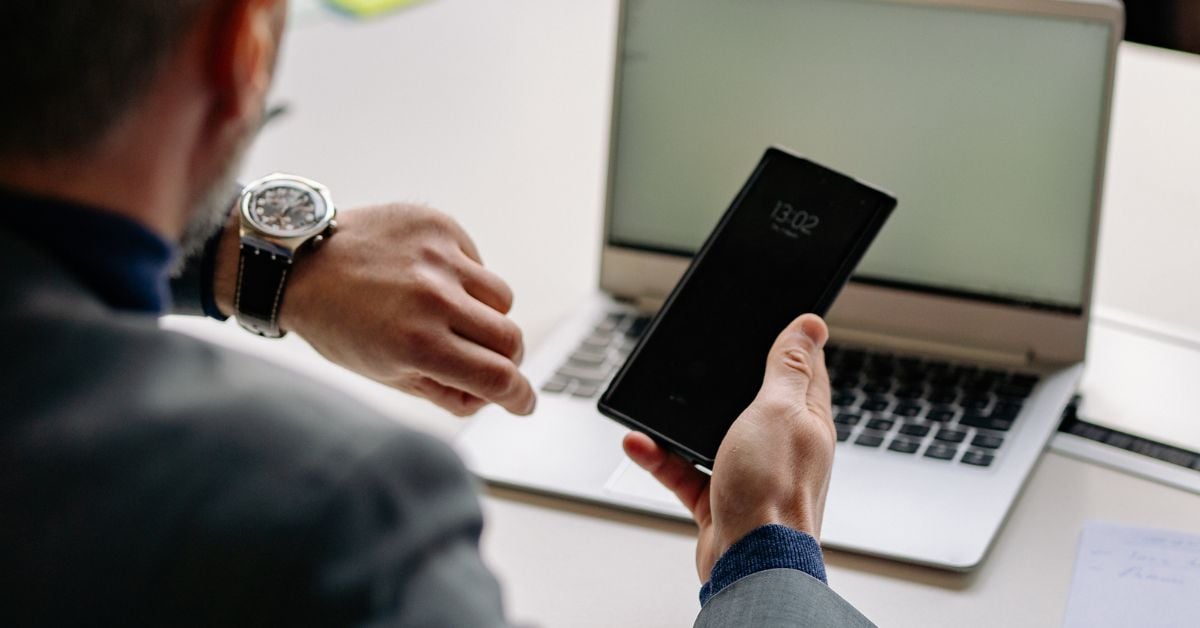 Persoon kijkt op horloge en houdt smartphone vast in andere hand terwijl hij aan een bureau zit met laptop
