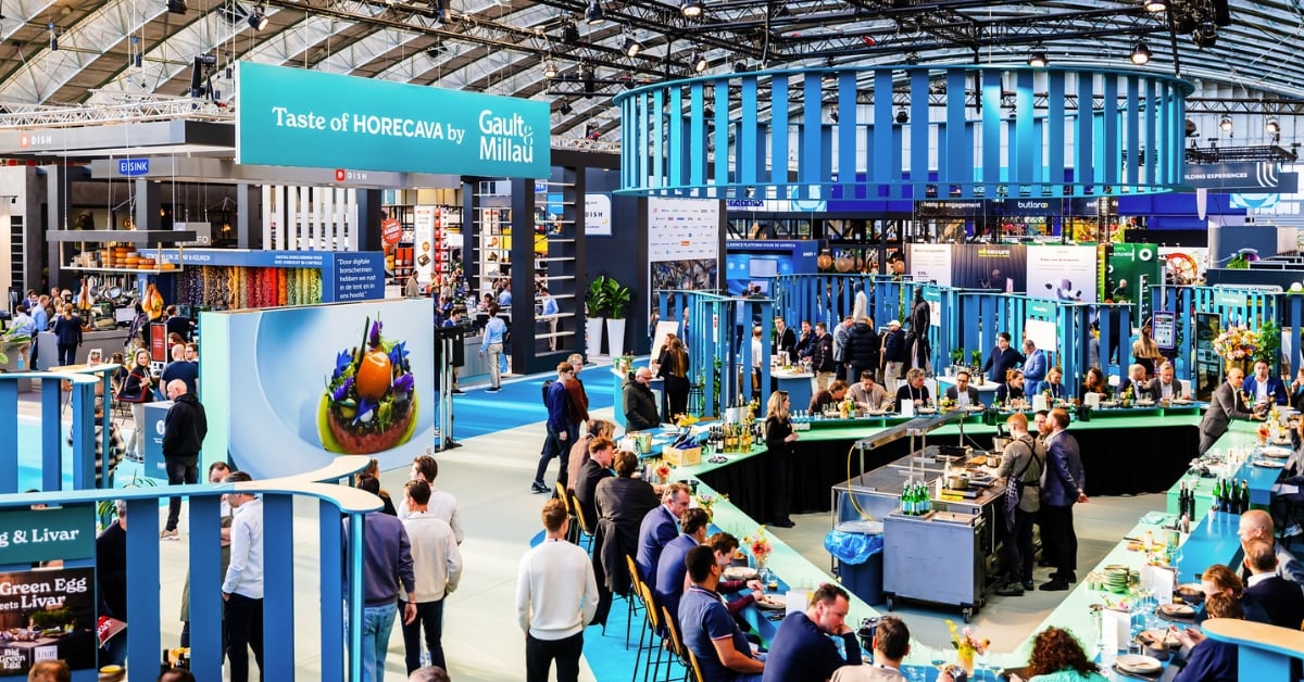 Beursvloer met mensen aan de chef's table, koks die live koken en verschillende stands met een bord “Taste of Horecava by Gault&Millau"