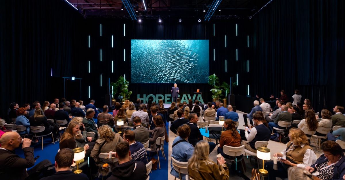 Bezoekers luisteren naar een spreker op het podium tijdens een presentatie, met een afbeelding van een school vissen op de achtergrond