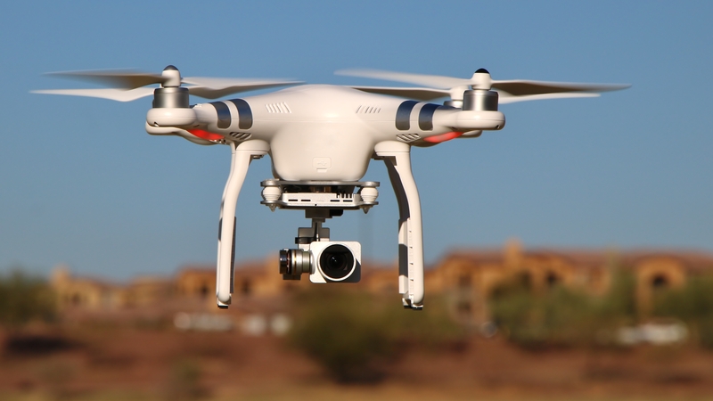 White quadcopter with gimbal camera hovering in clear skies above a suburban landscape.