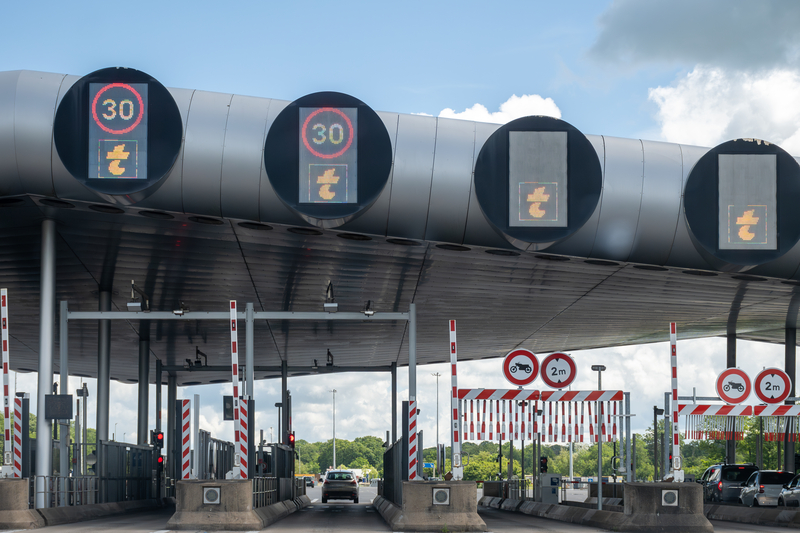 Toll plaza with electronic gantries showing 30 km/h and tag-only lanes, 2 m height limits and barriers as cars approach under a cloudy sky